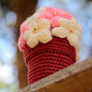 May include: A crocheted flower pot with pink and white flowers. The pot is a deep red colour and the flowers have gold centres.