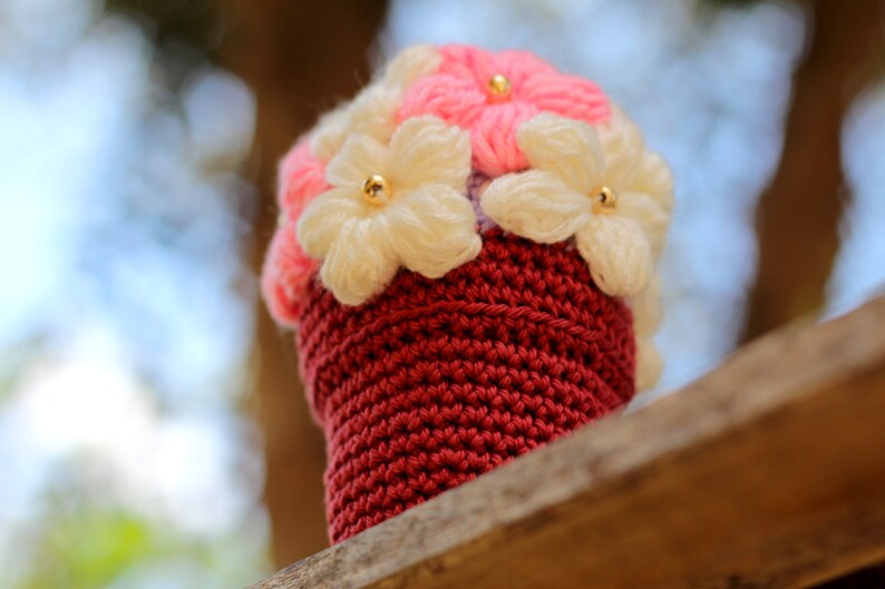 May include: A crocheted flower pot with pink and white flowers. The pot is a deep red colour and the flowers have gold centres.