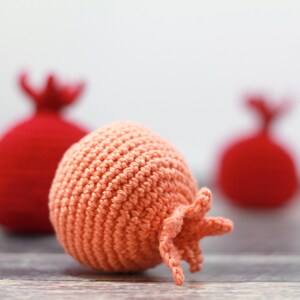 K&ouml;nnte beinhalten: Zwei rote geh&auml;kelte Granat&auml;pfel und ein pfirsichfarbener geh&auml;kelter Granatapfel. Der pfirsichfarbene Granatapfel ist scharf gestellt, die roten Granat&auml;pfel sind unscharf.