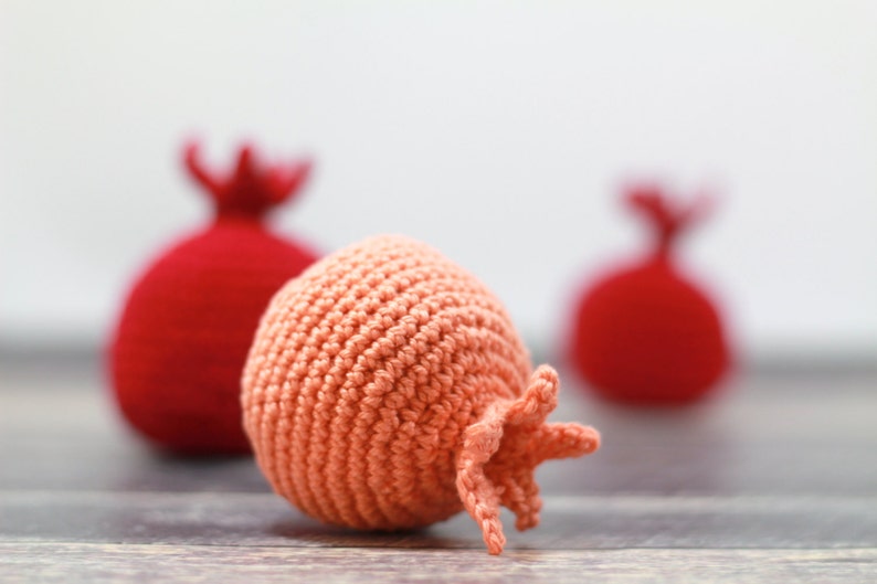 K&ouml;nnte beinhalten: Zwei rote geh&auml;kelte Granat&auml;pfel und ein pfirsichfarbener geh&auml;kelter Granatapfel. Der pfirsichfarbene Granatapfel ist scharf gestellt, die roten Granat&auml;pfel sind unscharf.