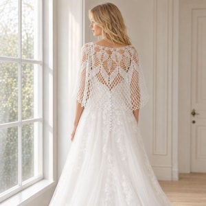 May include: A white crochet bolero featuring an intricate openwork diamond pattern and scalloped edges, worn over a floor-length white gown with a lace bodice and a vertical row of small round buttons running down the center of the back.
