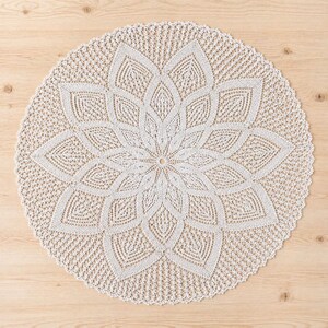 May include: A cream-coloured, circular doily with a floral design. The doily features a central flower pattern with detailed petal shapes and a scalloped edge. The intricate crochet work is visible throughout the piece, creating a delicate and textured appearance.