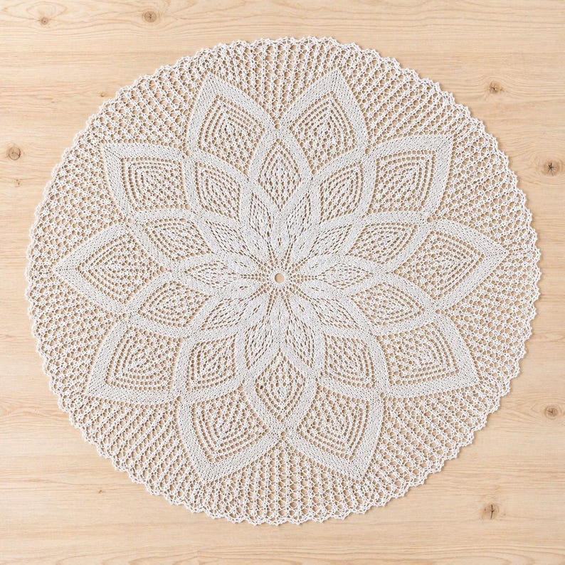 May include: A cream-coloured, circular doily with a floral design. The doily features a central flower pattern with detailed petal shapes and a scalloped edge. The intricate crochet work is visible throughout the piece, creating a delicate and textured appearance.