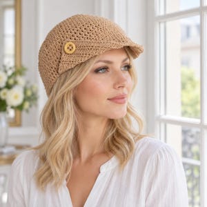 May include: A tan crochet newsboy cap featuring a textured, woven pattern and a small, round, light-colored wooden button detail on the side. The cap has a short, stiffened brim and is constructed from a light brown yarn material.
