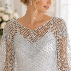 May include: A silver crocheted shawl with a V-neck design. The open weave of the shawl is visible, and it appears to be worn over a white garment. The overall style suggests a formal or bridal aesthetic.