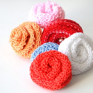 May include: Six crocheted flower embellishments in various colors, including pink, orange, red, blue, and white. The flowers are arranged in a cluster on a white background.