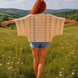 May include: A cream-coloured, short-sleeved, open-knit crochet top featuring vertical panels of intricate geometric patterns and a wide, boxy silhouette, paired with blue denim shorts that have a frayed, raw-edge hemline.