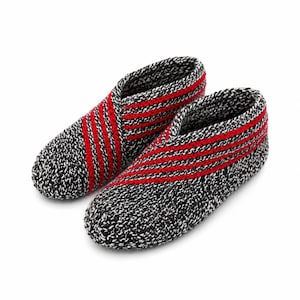 Könnte beinhalten: Ein Paar gestrickte Hausschuhe. Die Hausschuhe sind hauptsächlich schwarz und weiß mit roten Streifen auf der Oberseite. Sie bestehen aus dickem, strukturiertem Garn und wirken warm und bequem.