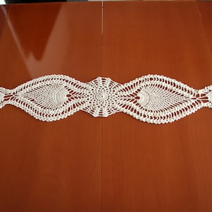 May include: White crocheted table runner with a delicate, intricate pattern. The runner is laid out on a wooden table.