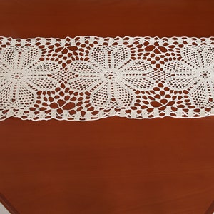 May include: A white crocheted table runner with a delicate floral pattern. The runner is laid out on a brown wooden table.