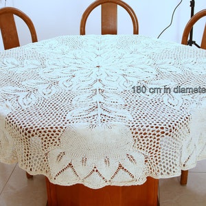 May include: A white crocheted tablecloth with a delicate floral pattern. The tablecloth is 180 inches in diameter and is laid out on a wooden table with three chairs.