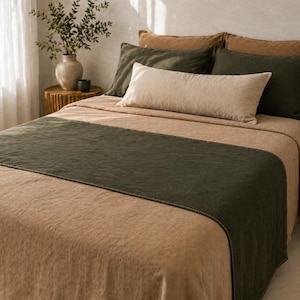 Könnte beinhalten: Ein Bett mit einer beigefarbenen Bettdecke und einem dunkelgrünen Bettläufer. Das Bett ist mit mehreren Kissen in Beige- und Dunkelgrüntönen ausgestattet. Eine Vase mit Grün steht auf einem hölzernen Beistelltisch.