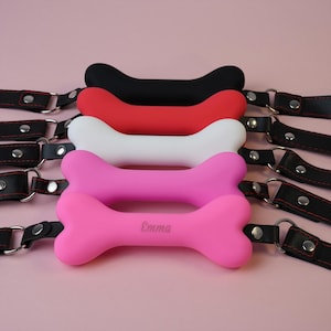 Peut inclure: Cinq bâillons en forme d'os de couleurs noir, rouge, blanc, rose clair et rose vif. Chaque bâillon est fixé à des lanières en cuir noir avec des coutures rouges et des ferrures métalliques argentées. Le bâillon rose vif porte le nom Emma gravé.