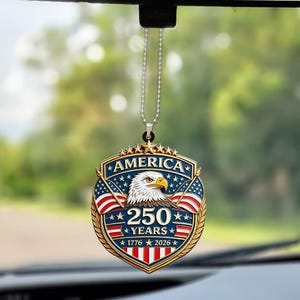 May include: A patriotic car ornament featuring a shield design with an eagle, American flags, and the text "AMERICA 250 YEARS 1776 2026". The ornament is suspended by a silver chain and hangs from a car's rearview mirror.
