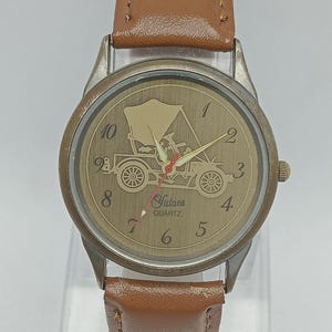 Op de afbeelding: Een vintage horloge met een bruine leren band. De wijzerplaat toont een goudkleurig antiek auto-ontwerp met het opschrift "Futaca Quartz". Het horloge heeft een zilverkleurige metalen behuizing.