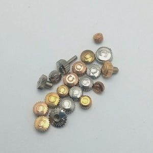 Peut inclure: Un assortiment de couronnes de montre dans diverses finitions métalliques, argent, or et bronze. Les couronnes présentent un design circulaire avec un logo au centre. Certaines couronnes ont un bord texturé et strié. L'assortiment est dispersé sur une surface blanche.