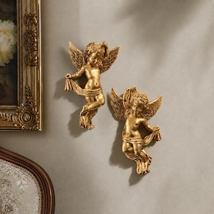 May include: Two gold-coloured wall-mounted cherub sculptures, each featuring a winged figure with curly hair draped in a flowing cloth. The cherubs are depicted in a dynamic, mid-air pose with detailed feathers on their wings and smooth, metallic finishes.
