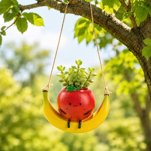 May include: A hanging planter shaped like a red apple with a smiling face, resting on a yellow banana-shaped base. The apple contains a small green succulent plant. The entire piece is suspended by thin brown twine from a tree branch.