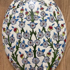 Op de afbeelding: Ovale keramische schaal met een bloemmotief. Het ontwerp toont blauwe, rode en gele bloemen met groene stelen en bladeren tegen een witte achtergrond. De schaal staat op een houten oppervlak.