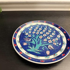 Puede incluir: Un plato redondo de cerámica pintado a mano con un diseño floral azul y blanco. El plato presenta un motivo central de árbol con flores blancas y detalles en rojo. El borde exterior tiene un patrón repetitivo de elementos florales rojos y azules.