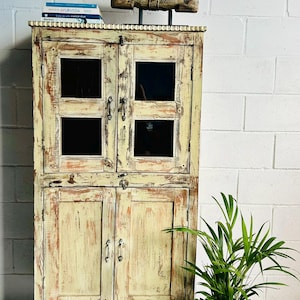 Può includere: Un mobile in stile vintage bianco sporco con quattro ante a vetri e maniglie in metallo. Una scultura di cavallo in legno intagliato è posta sopra, con dei libri. Una pianta in vaso in un cesto intrecciato è a destra. Il mobile è largo circa 100 cm.