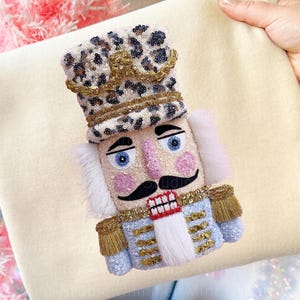 May include: A decorative nutcracker figurine with a leopard print hat and blue eyes. The figurine has a black moustache, pink cheeks, and a blue and gold uniform. The nutcracker is embellished with sequins and gold trim.