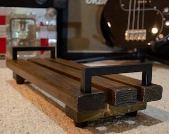 May include: A wooden pedalboard with a dark finish and black metal handles. The pedalboard is rectangular and has a simple, functional design. It is likely used for musical instruments, such as guitars or effects pedals.