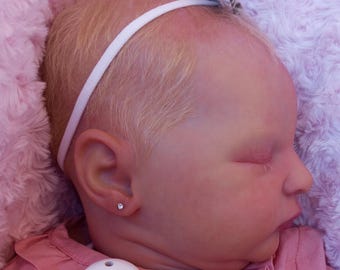 May include: A realistic baby doll with closed eyes, dressed in a pink outfit and a white headband with a bow. The doll has a small diamond earring and a dummy clip. The doll is resting on a pink, fluffy blanket.