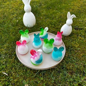 Könnte beinhalten: Zwei große weiße Osterhasenfiguren und ein Tablett mit bunten seifenförmigen Hasen. Die Seifen sind in Rosa-, Grün- und Blautönen gehalten. Der Hintergrund ist grünes Gras.