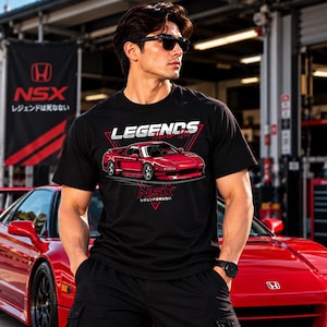 May include: A black short-sleeved t-shirt featuring a graphic of a red Honda NSX sports car. The design includes the white text "LEGENDS Never Die" above the car and the red text "NSX" with Japanese characters below the car on a red triangle.
