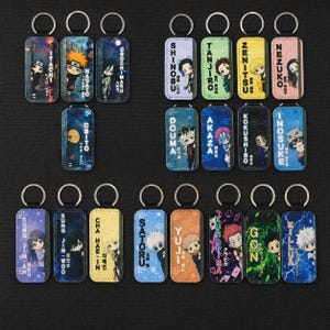 May include: A selection of rectangular keyrings with anime character designs. Each keyring has a metal loop and a printed image with character names such as "Itachi", "Naruto", and "Killua". The backgrounds are various colours, including blue, yellow, and orange.