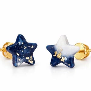 Könnte beinhalten: Ein Paar sternförmige Ohrstecker. Jeder Ohrring hat einen goldenen Stecker und einen Stern mit einem dunkelblauen und weißen Marmoreffekt, der mit goldenen Flocken übersät ist. Die Ohrringe sind vor einem weißen Hintergrund.