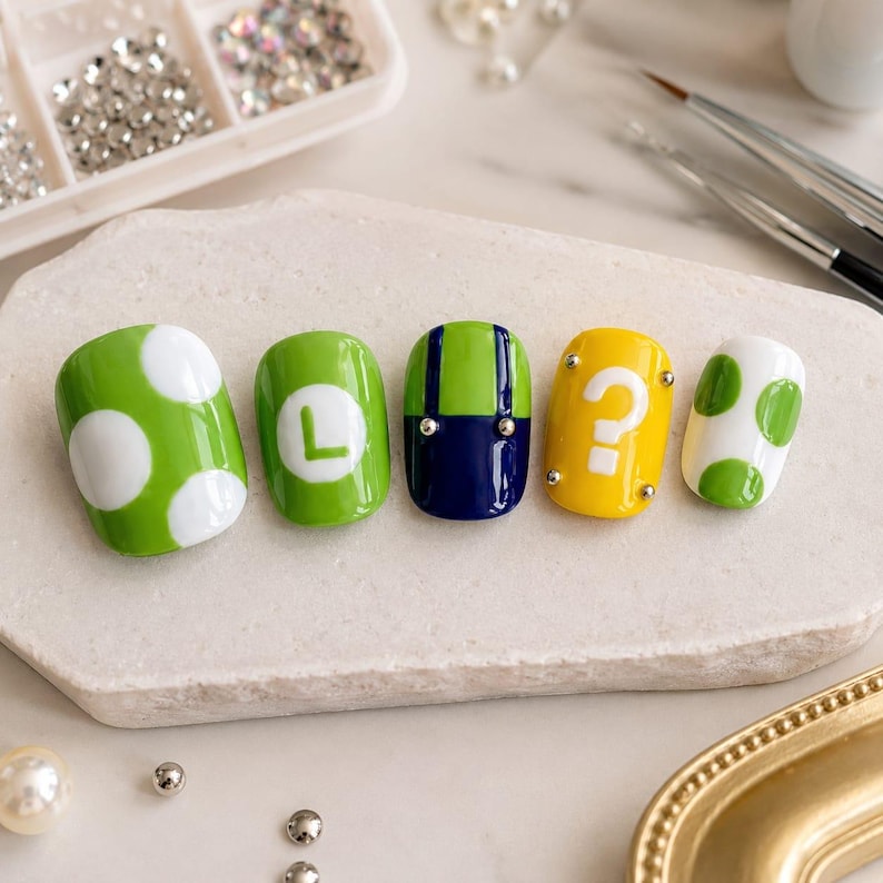 May include: Five press-on nails featuring vibrant designs: a green nail with white polka dots, a green nail with a white letter L, a green and navy blue nail resembling overalls, a yellow nail with a white question mark, and a white nail with green dots.