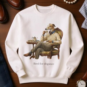 Puede incluir: Sudadera blanca con una ilustración de un galgo sentado, bebiendo té. La imagen incluye un reloj, zapatos, vaqueros y una pulsera de perlas. El texto "Bred for elegance" está impreso debajo.