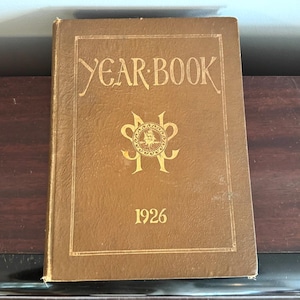 May include: A vintage yearbook from 1926. The cover is a textured brown with gold lettering that reads "YEAR BOOK" and "1926". A gold emblem is centered on the cover.