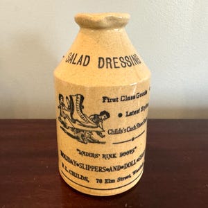 May include: A vintage, tan-colored ceramic bottle with the words "SALAD DRESSING" in black lettering. The bottle features a black and white illustration of ice skates and text that reads "LADIES' RINK BOOTS".