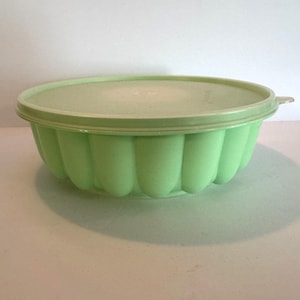 May include: A light green, vintage-style gelatin mold with a matching lid. The mold has a fluted design around the sides and a round shape. The lid has a small handle. This kitchenware item is perfect for making desserts.