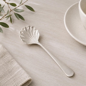 May include: A silver-toned metal spoon featuring a bowl shaped like a scalloped seashell with detailed ridges radiating from the center to the edge. The handle is smooth, slender, and tapers slightly toward the rounded end of the utensil.