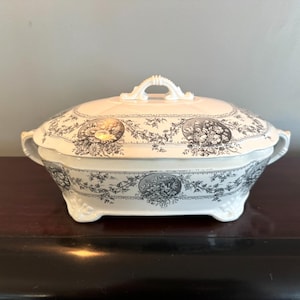 May include: A vintage white and grey ceramic serving dish with a lid. The dish has a floral design and a handle on the lid. The dish has a footed base and handles on the sides.