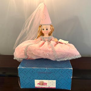 Könnte beinhalten: Eine Vintage-Puppe in einem rosa Kleid und passendem konischen Hut, verziert mit silbernen Verzierungen und Pailletten. Die Puppe hat blondes Haar und einen durchsichtigen Schleier. Sie sitzt auf einer blauen Schachtel mit dem Etikett "Fairy Godmother".