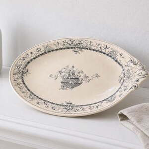 May include: An oval ceramic platter with a cream-colored base and a dark gray floral transferware pattern. The design features a central bouquet in a decorative urn and a detailed botanical border with a scalloped edge on one side.