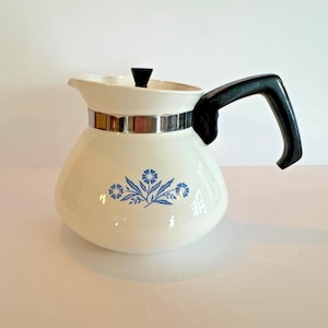 Puede incluir: Cafetera vintage de color crema con asa y tapa negras. La cafetera presenta una banda plateada y un diseño floral azul. El pico vertedor está en el lado izquierdo. Aproximadamente 20 cm de altura.