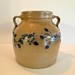May include: A tan stoneware crock with two handles and a blue floral design. The jar has a cylindrical shape with a slightly wider base and a narrow neck. The blue floral design includes leaves and berries.