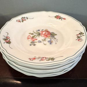 May include: A stack of white ceramic soup bowls with a scalloped edge and a green trim. Each bowl features a floral design in the center and around the rim, with pink, red, and blue flowers. The bowls are stacked on a dark surface.