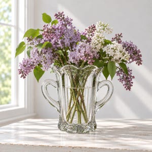 May include: A clear glass vase with two side handles and a scalloped rim, filled with a large bouquet of fresh purple and white lilac flowers with green leaves. The vase features vertical ribbed detailing and a textured, wavy base design.