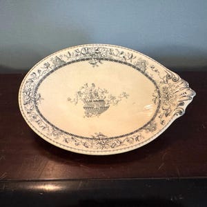 May include: Oval serving platter with a cream-colored surface and a blue floral design. The platter features a central floral arrangement and a decorative border. The handle is on the right side.