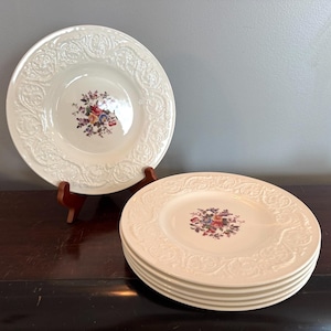 Könnte beinhalten: Ein Satz cremefarbener Keramikteller mit floralem Mitteldesign. Die Teller haben einen dekorativen, geprägten Rand. Ein Teller steht auf einem Holzständer, die anderen sind auf einer dunklen Holzoberfläche gestapelt.