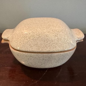 May include: A speckled ceramic lidded dish with a rounded shape. The off-white dish has small dark flecks and a light brown rim. It features two small handles on either side.