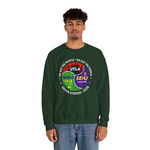 May include: A dark green sweatshirt with a circular graphic featuring the text "WE GOT THE PEOPLE" and "WE GOT THE POWER". The graphic includes the words "UTLA", "TEAMSTERS AALA Local 2010", and "SEIU Local 99". The words "UNION STRONG 2026" are also present.