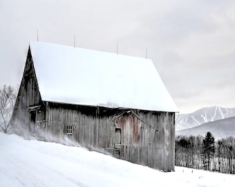 May include: A weathered wooden barn with a snow-covered roof stands in a snowy field. The barn has a red door and is surrounded by trees. In the background, a mountain range with ski slopes is visible.